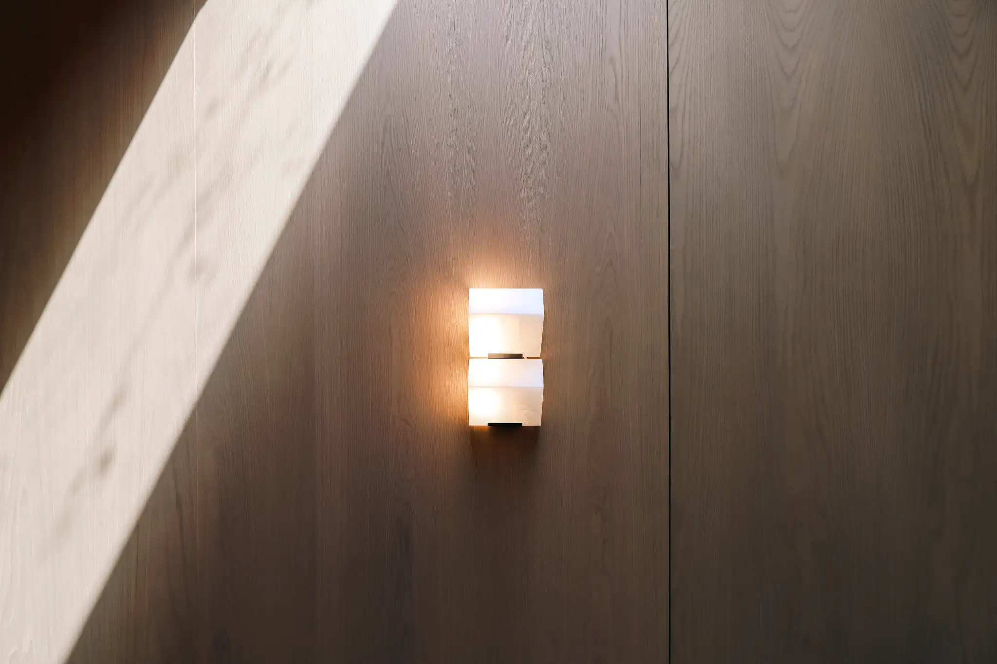 Modern white wall sconce mounted on light wood panel wall with sunlight casting shadows.