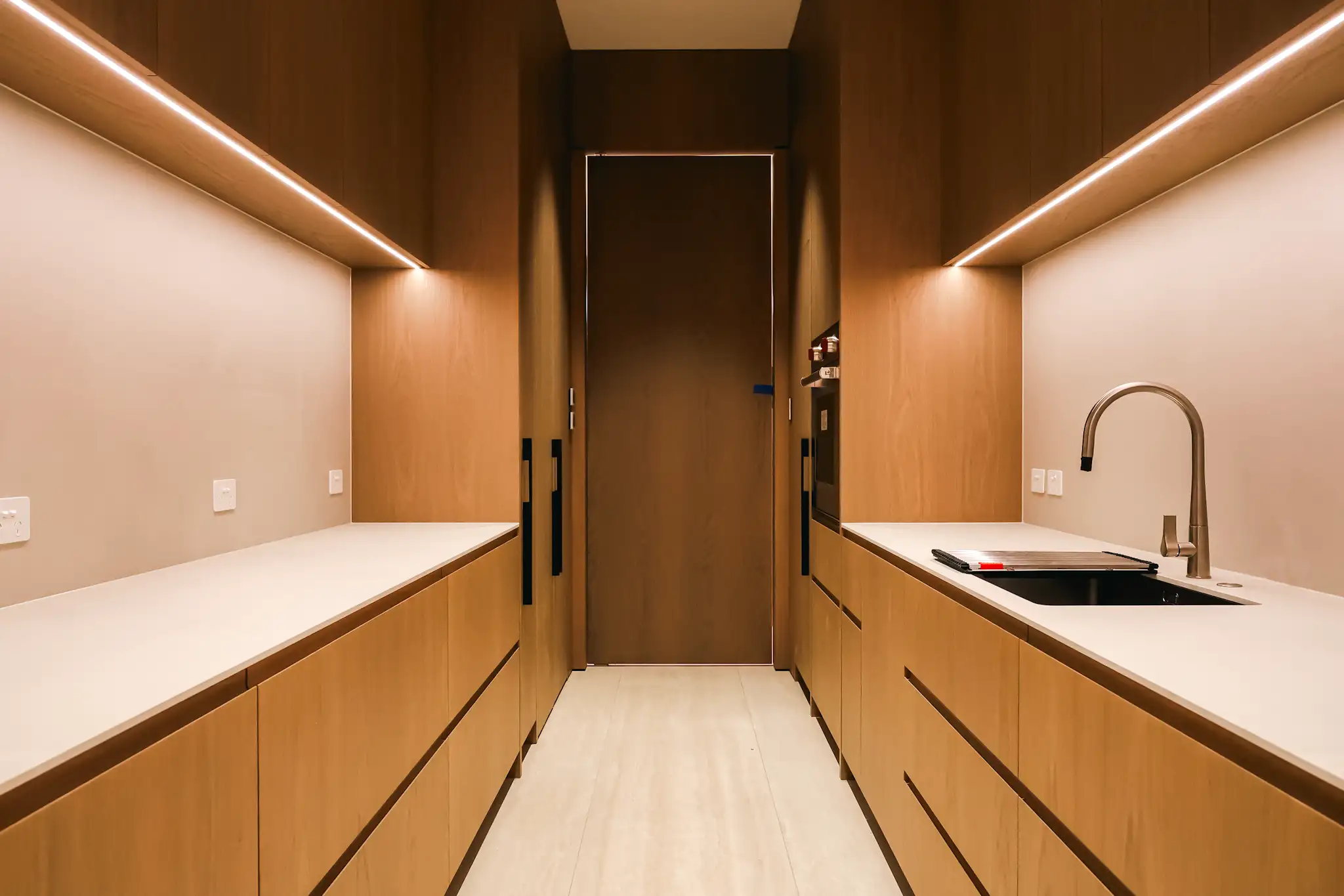 Minimalist kitchen with wood cabinetry, white countertops, under-cabinet lighting, a built-in oven, and a stainless steel faucet over a sink.