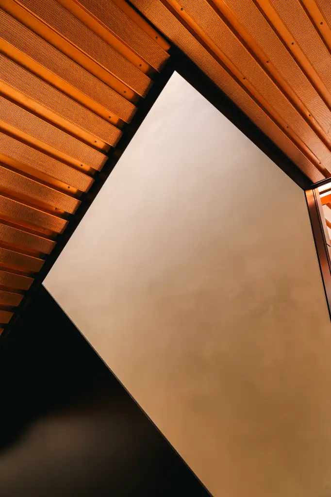 Abstract architectural detail showing an upward view of a geometric ceiling with orange perforated panels and a large central triangular skylight.