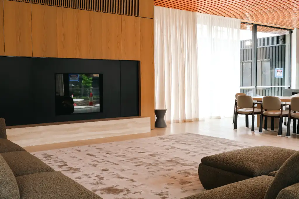 Modern living room with brown couches, a patterned rug, a black fireplace with wooden paneling, and a dining area near large windows with sheer white curtains.