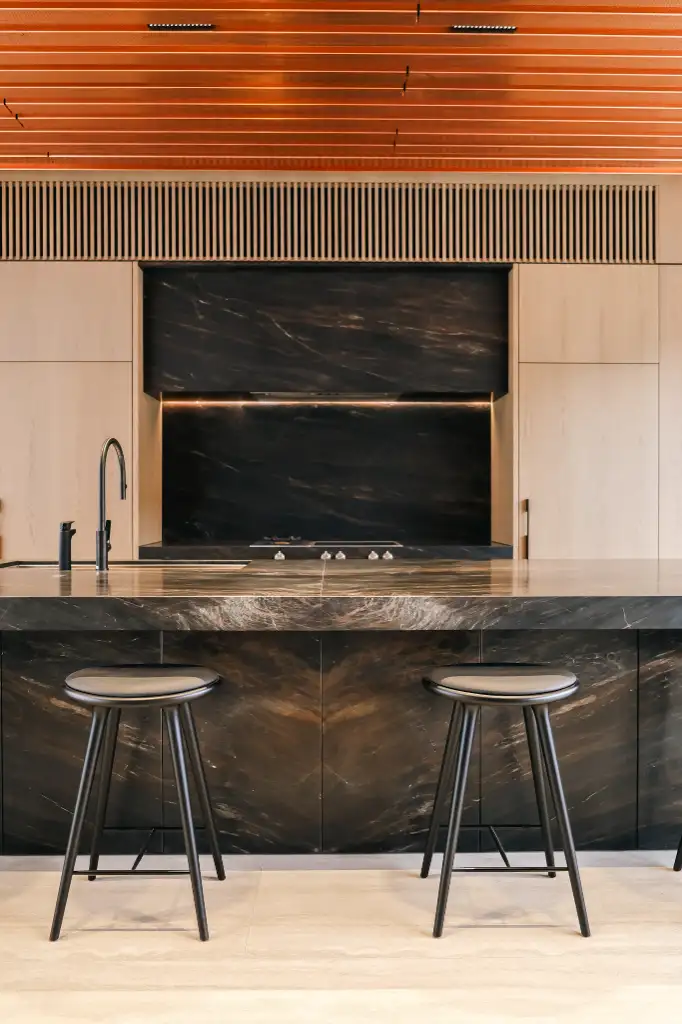 Modern kitchen with a black marble island, two black stools, light wood cabinets, and a sleek black faucet.