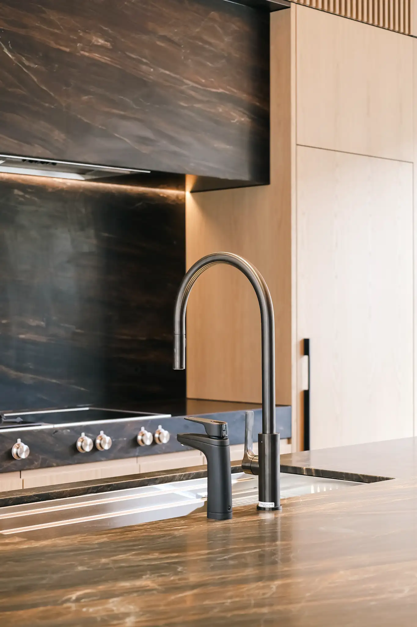 Modern kitchen sink with a tall black curved faucet and a shorter black tap on a brown marble countertop.