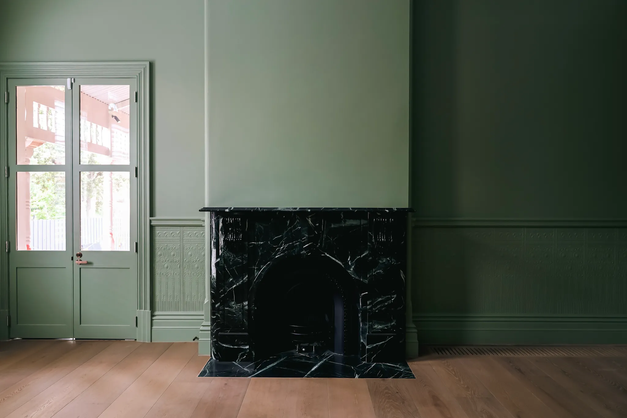 Empty room with green walls, a black marble fireplace, and a wooden floor next to glass double doors.