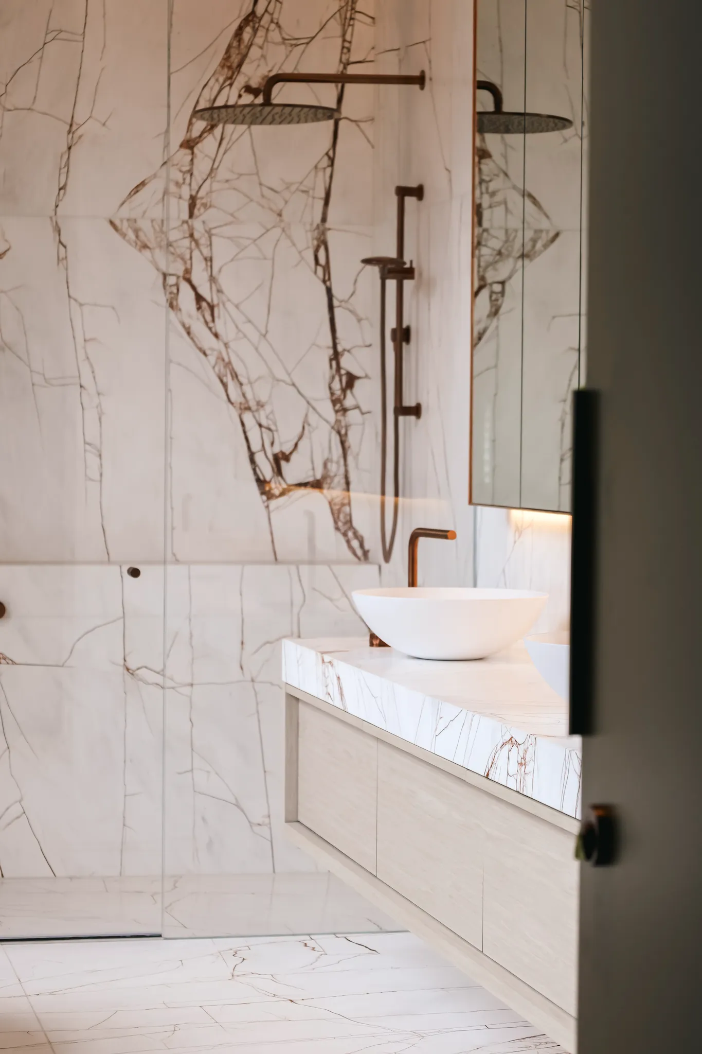 Modern bathroom with marble walls and floor, a glass shower with a rain showerhead, and a white vessel sink on a marble countertop with bronze fixtures.