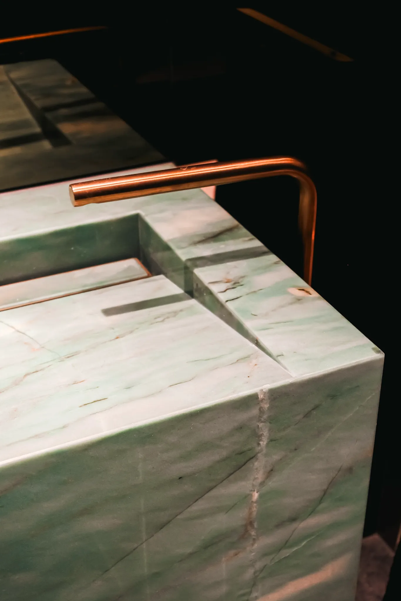 Modern rectangular marble sink with greenish hues and a sleek copper faucet in a dimly lit setting.
