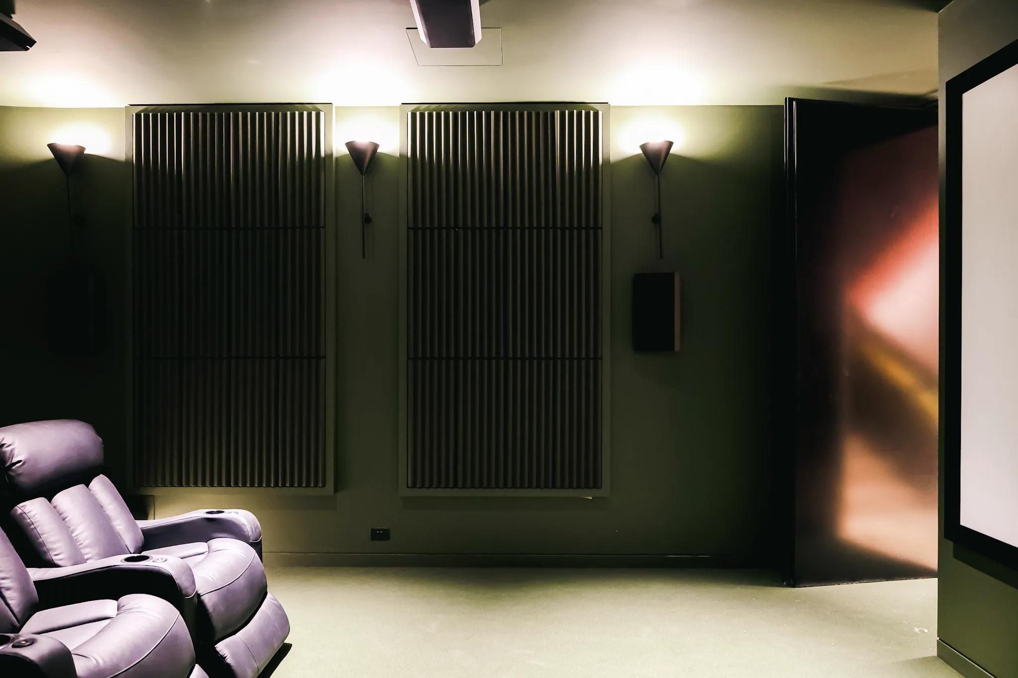 Dark home theater room with leather recliner seats, wall-mounted acoustic panels, and soft wall lights.