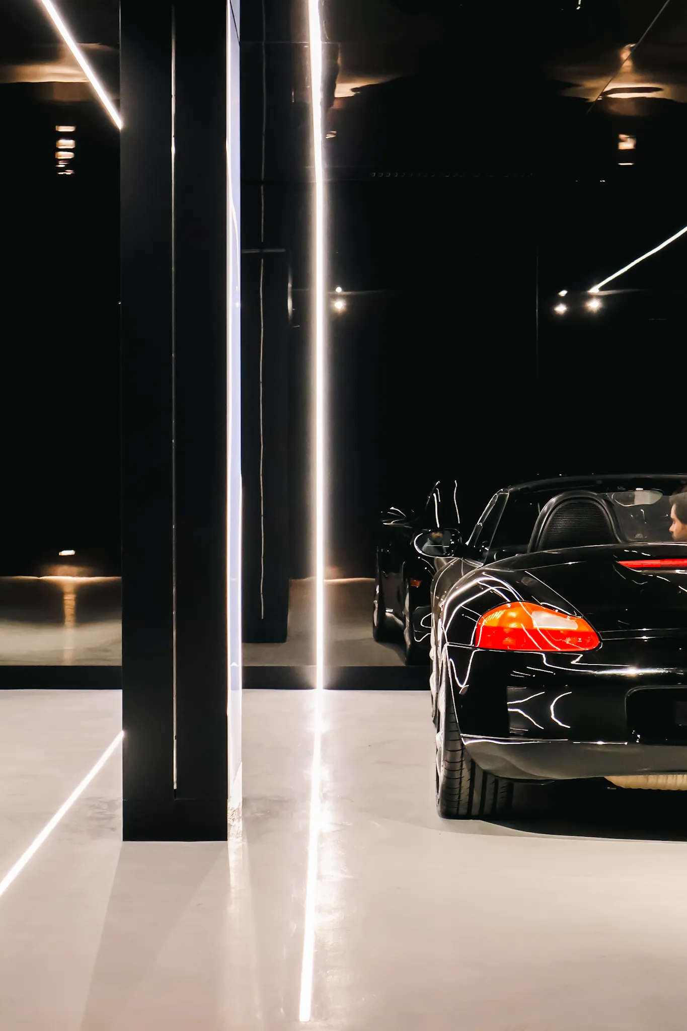 Rear view of a black convertible sports car parked indoors beside black reflective panels with vertical white light strips.
