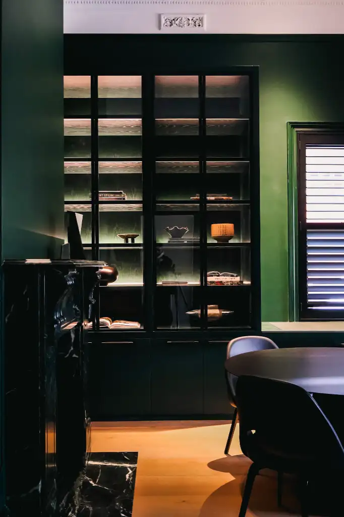 Dark green wall with a black glass-front cabinet displaying decorative items, next to a window with shutters and a black fireplace to the left.