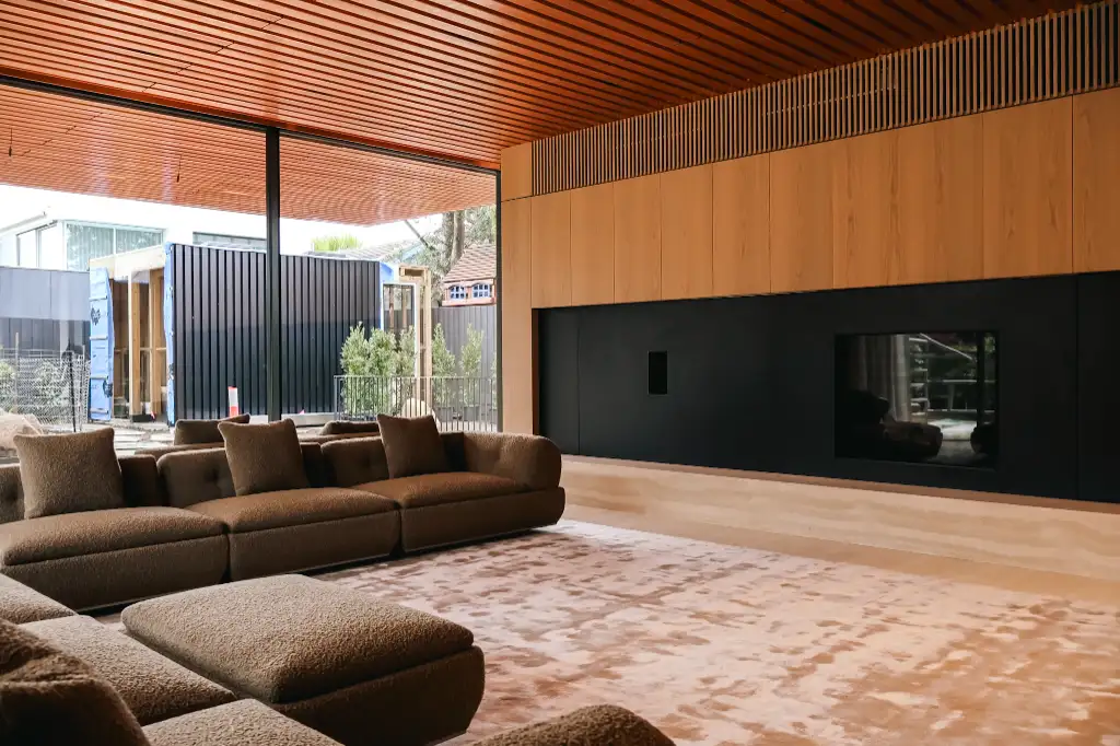 Modern living room with brown sectional sofa, beige patterned carpet, wood-paneled walls, and a large window showing outdoor containers.