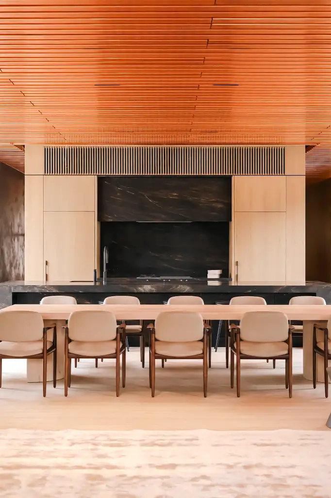 Modern dining area with light wood table and beige chairs in front of a black marble kitchen counter under a wooden slatted ceiling.